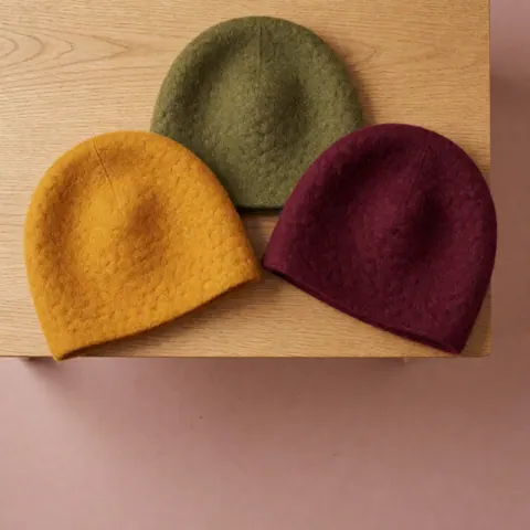 Three freshly dyed wool hats in warm tones arranged in a triangle on a light wood table against a dusty pink wall.