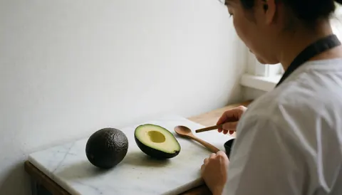 A chef prepares a fresh avocado on a white marble table with a wooden spoon and soft natural light