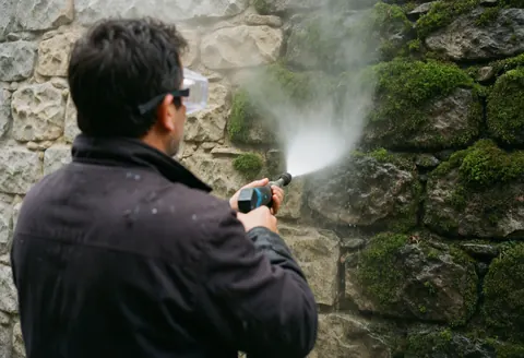 Worker wearing safety goggles uses a water pressure gun to clean moss off a stone wall, showing the contrast between dirty and clean surfaces in a realistic landscaping scene.