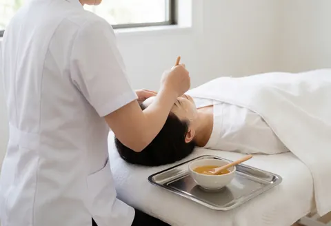 A therapist applies a honey mask to a client's face in a clean, professional aesthetic clinic setting with a treatment bed and tray.