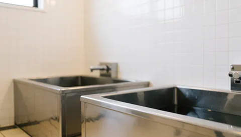 Empty stainless steel dog grooming tub with faucet and drain, set against a clean white tiled wall, ideal for text background