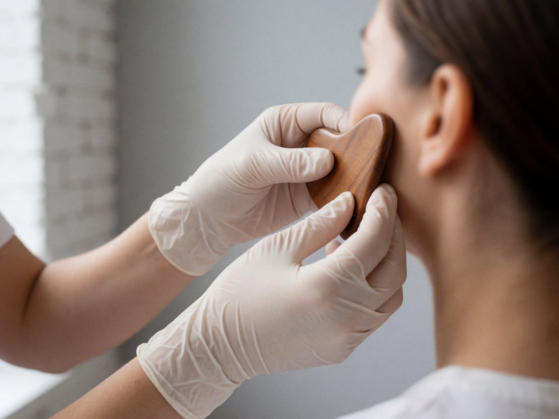 Gloved hands performing a gua sha facial treatment on a cheek against a gray background with a brick wall, showcasing professional skincare care.