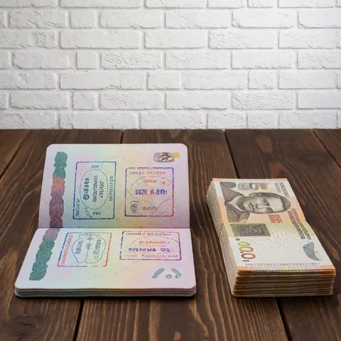 Open passport displaying travel stamps alongside a stack of local banknotes resting on a dark wooden table surface