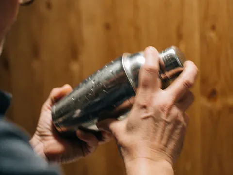 Close-up of hands shaking a metal cocktail shaker with condensation against a warm wooden background