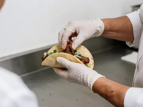 Close-up view of gloved hands preparing a fresh taco with grilled meat, cilantro, and onion on a stainless steel surface for fast food service