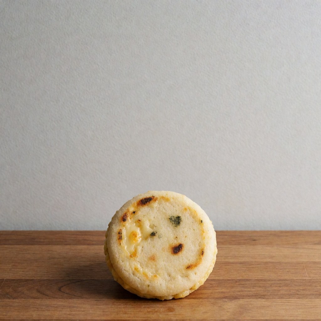 Top view of a small arepa with melted cheese on a wooden surface against a light gray wall, representing fast food.