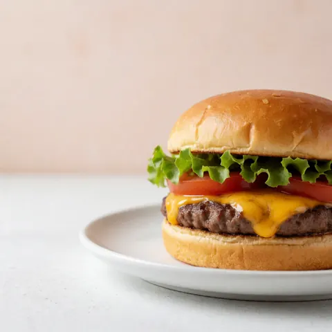 Classic cheeseburger with melted cheese, lettuce, and tomato served on a white plate against a pale terracotta wall with natural lighting.