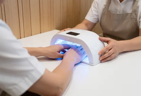 Professional manicurist using a white UV lamp to cure gel nails on a client in a clean, modern salon with light wood walls.