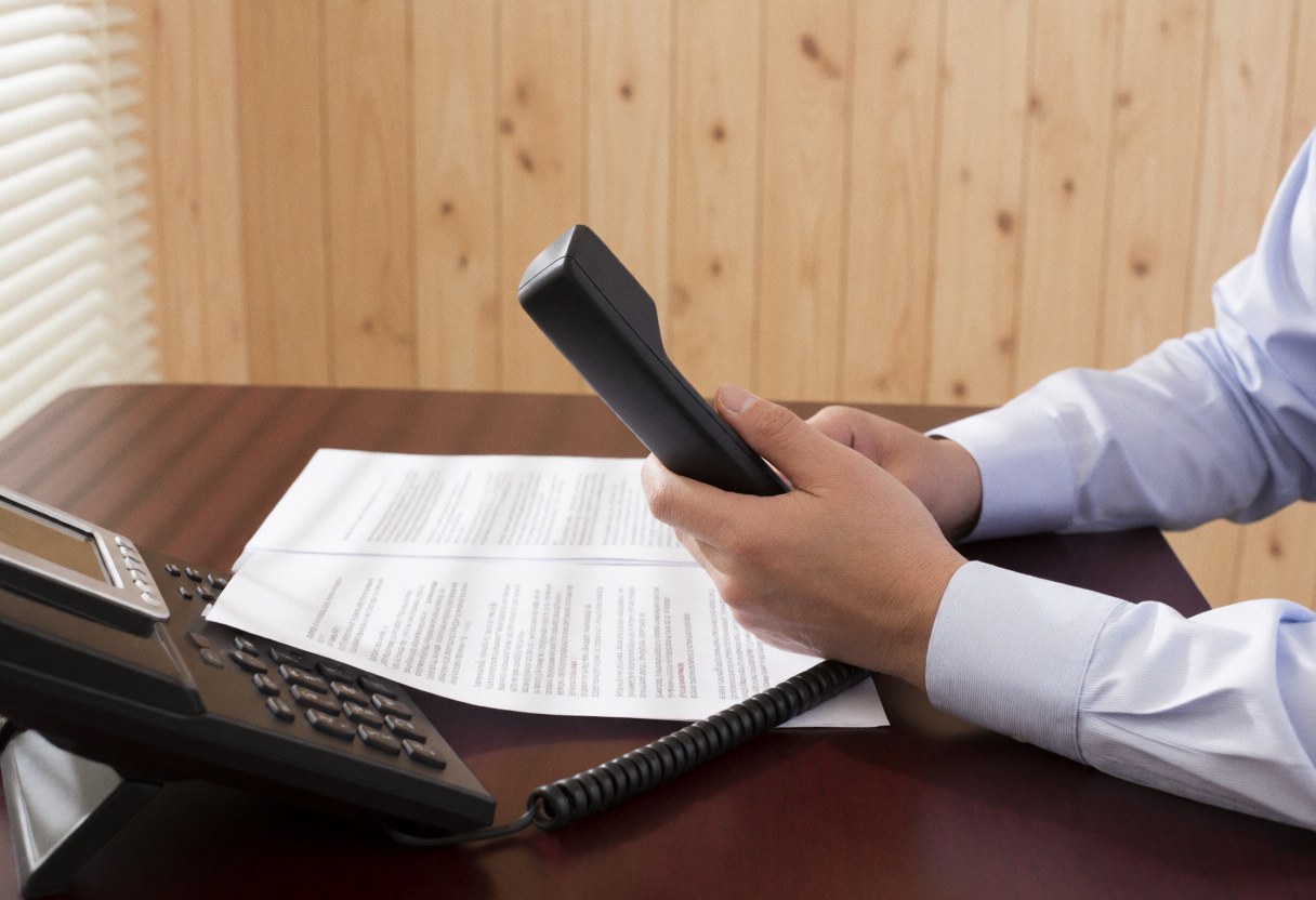 A professional lawyer sits at a wooden desk in an office, holding a phone receiver while reviewing important documents during a serious business negotiation.