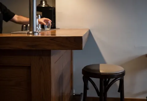 Polished wooden bar counter with a bar stool in the foreground, gray wall background, bartender hand visible, warm atmospheric lighting.