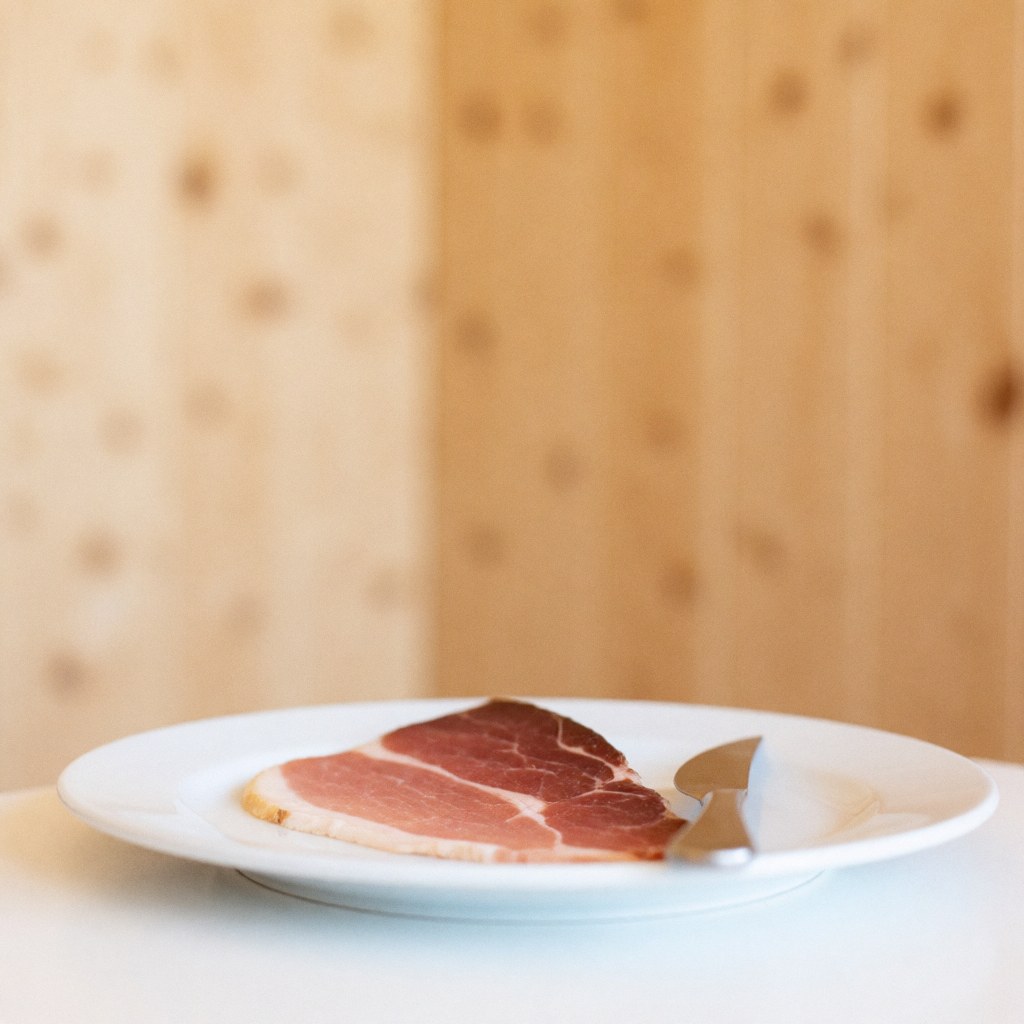 A single slice of cured ham served on a white plate with a knife on a wooden surface, ideal for bar and cocktail product imagery