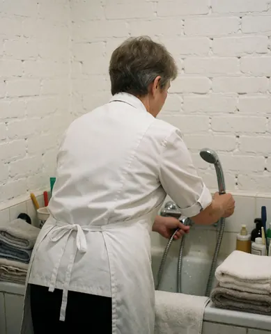 A groomer adjusts a showerhead over a tub in a white brick room with neatly folded towels, showcasing a clean and organized pet care environment.