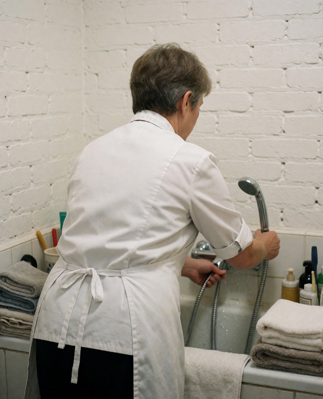 A groomer adjusts a showerhead over a tub in a white brick room with neatly folded towels, showcasing a clean and organized pet care environment.