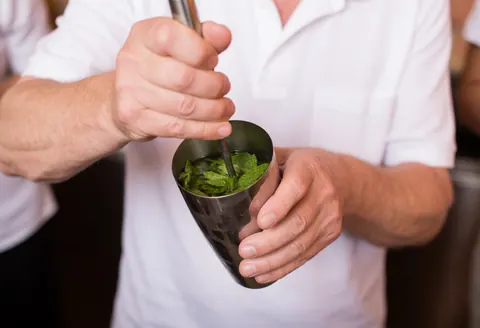 Professional bartender muddling fresh mint leaves in a metal shaker with a blurred terracotta wall background