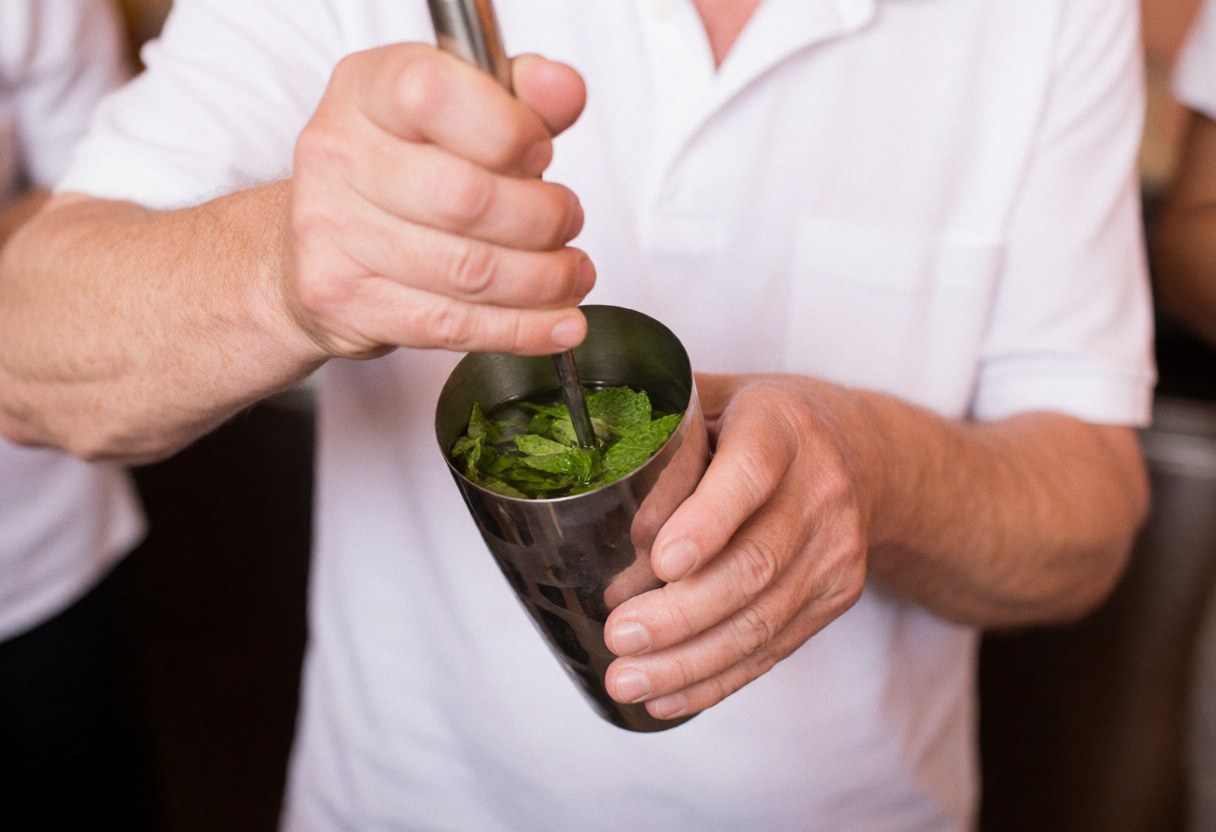Professional bartender muddling fresh mint leaves in a metal shaker with a blurred terracotta wall background