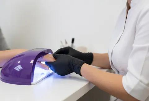 Close-up of a manicurist's gloved hand holding a client's finger under a uv lamp for gel nail curing