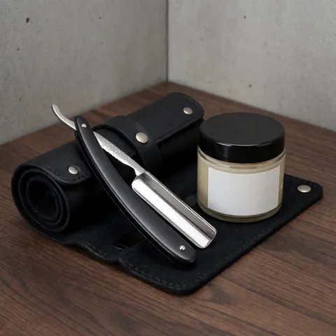 Straight razor resting on a black leather case beside a shaving soap jar on a dark wood surface against a concrete wall