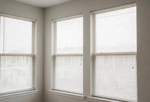 Empty and clean living room with three large windows covered by open white blinds, light gray walls, soft natural lighting, and a tidy atmosphere ideal for home cleaning services.
