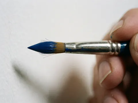 Macro detail of a blue paintbrush held by a hand, showing wet paint and visible bristles against a soft white background
