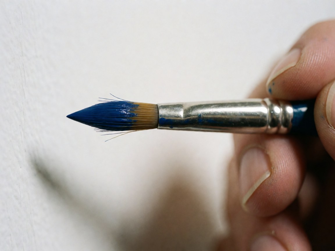 Macro detail of a blue paintbrush held by a hand, showing wet paint and visible bristles against a soft white background