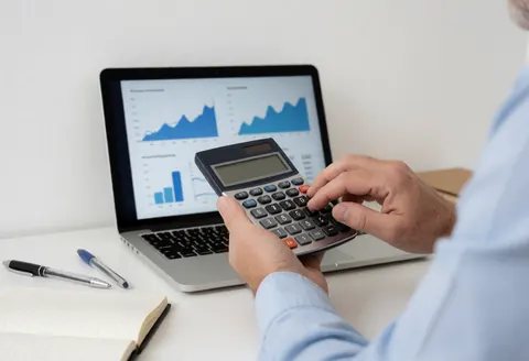 Business consultant checks numbers on a calculator while working on a laptop with financial charts on a clean desk with a notebook and pens.