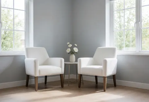 Empty treatment room featuring two white chairs, a table with flowers, large windows, gray walls, and light wood flooring.