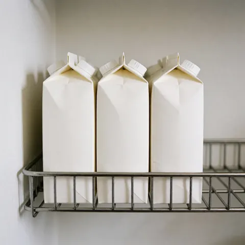 Three plain white milk cartons arranged on a metal shelf against a white wall with soft lighting, suitable for grocery store displays.