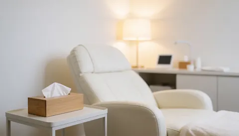 A clean white spa room featuring a reclining treatment chair, a wooden side table with tissues, and soft lighting for a calm atmosphere