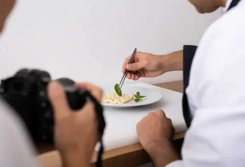 A food stylist uses tweezers to place a herb leaf on a dish while a photographer captures the scene in a bright kitchen studio.