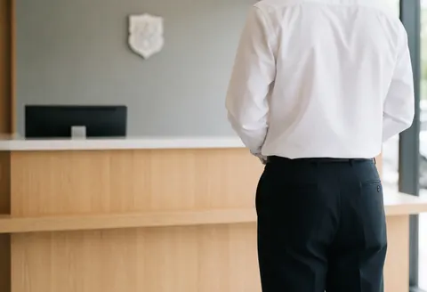 Person standing with back to camera at a modern reception area featuring a light wood desk, black monitor, and gray wall in a professional setting