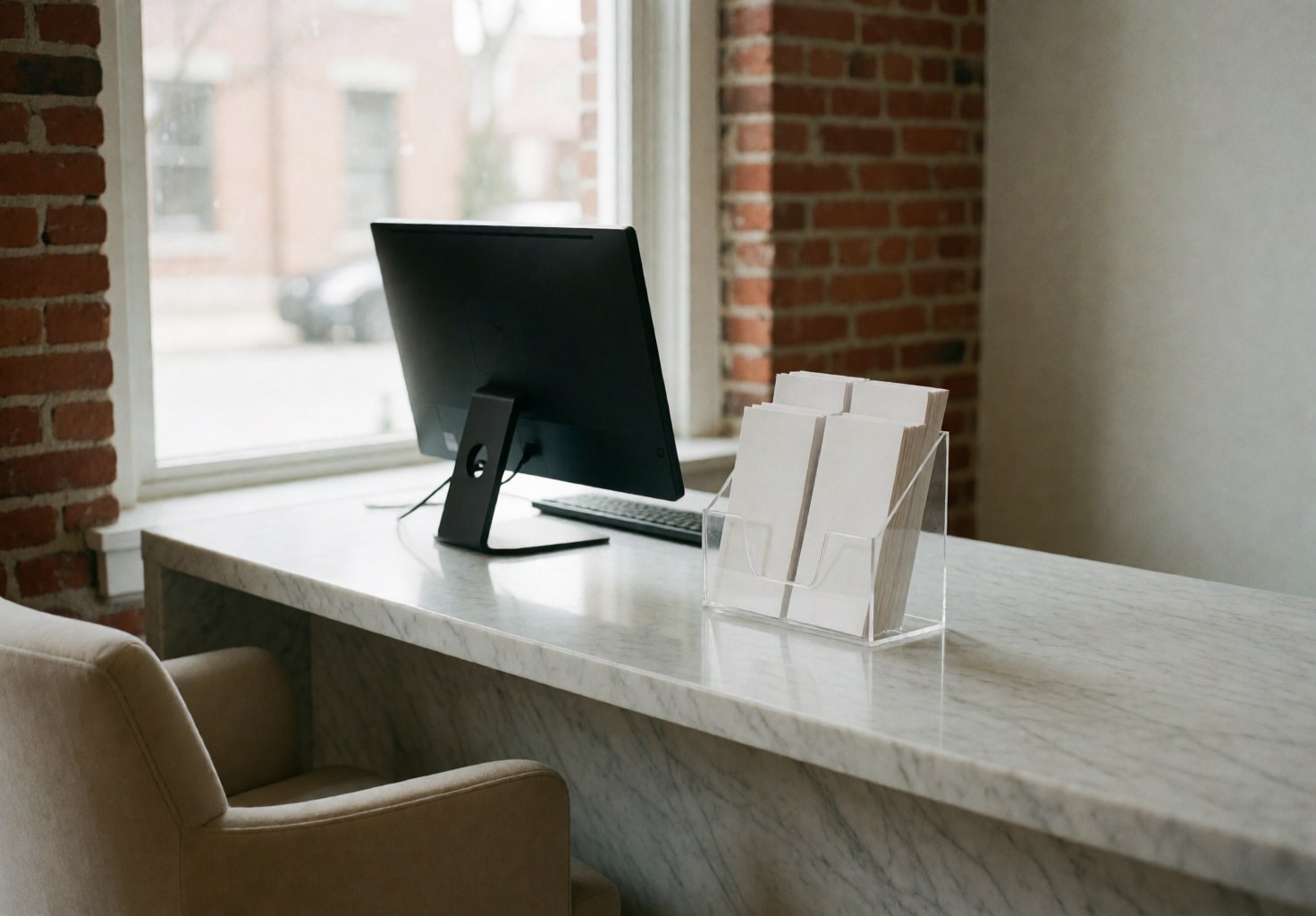 Minimalist spa reception area featuring a marble desk, computer, chair, and exposed brick wall, creating a calm and welcoming atmosphere for clients.