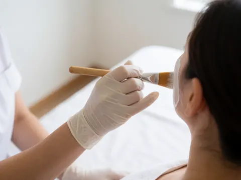 A professional wearing white gloves uses a wooden brush to apply a facial mask to a client against a white background with natural light