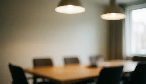 A warm, blurred background of a real estate office interior featuring a wooden table, chairs, and soft lighting near a window with curtains, ideal for text overlay.