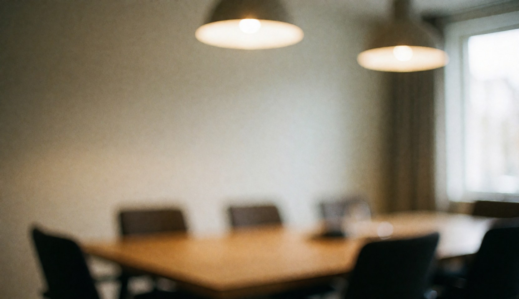 A warm, blurred background of a real estate office interior featuring a wooden table, chairs, and soft lighting near a window with curtains, ideal for text overlay.