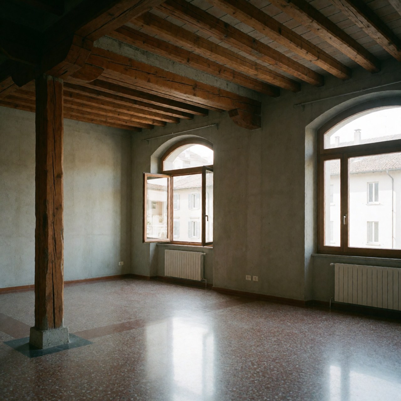 Interior view of a restored living room featuring exposed wooden beams, terrazzo flooring, arched windows, and concrete walls with natural light