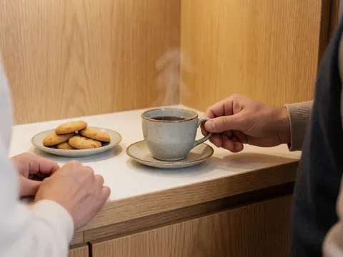 A hand holds a steaming coffee cup next to a plate of cookies on a white countertop with light wood walls.