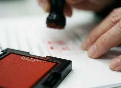 Close-up of a hand using a red ink stamp on a white paper document with a blurred background, suitable for accounting and office contexts.