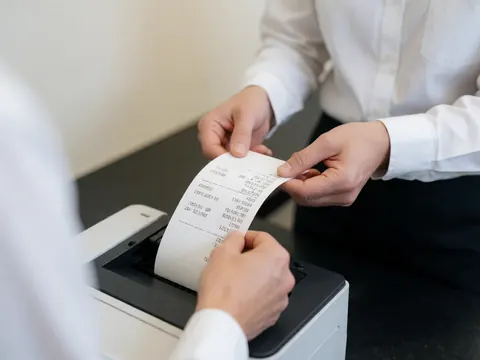 Close-up view of a printed thermal receipt held in a hand, showing transaction details against a dark background with white wall and clothing elements.