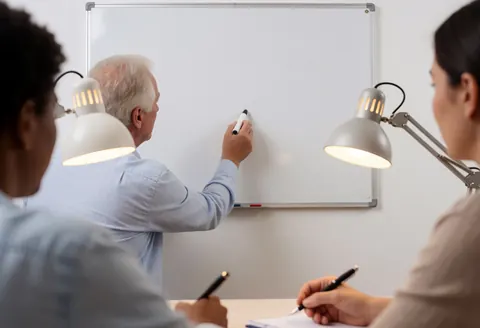 Tutor writes on a whiteboard while two students take notes with pens under desk lamps in a realistic language class setting.