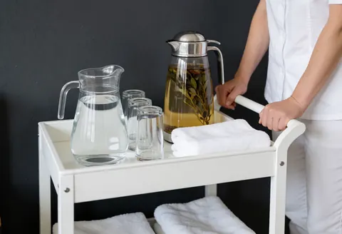 White spa cart holding water, tea, and towels against a gray charcoal wall, ideal for spa and massage interior decor