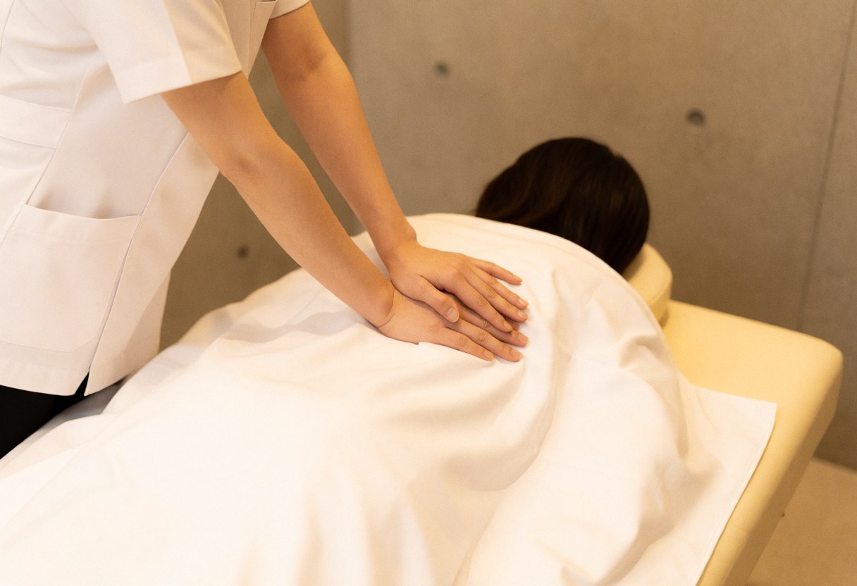 Therapist in white uniform applying therapeutic back massage to client covered with white sheet on massage table in serene room with polished concrete walls