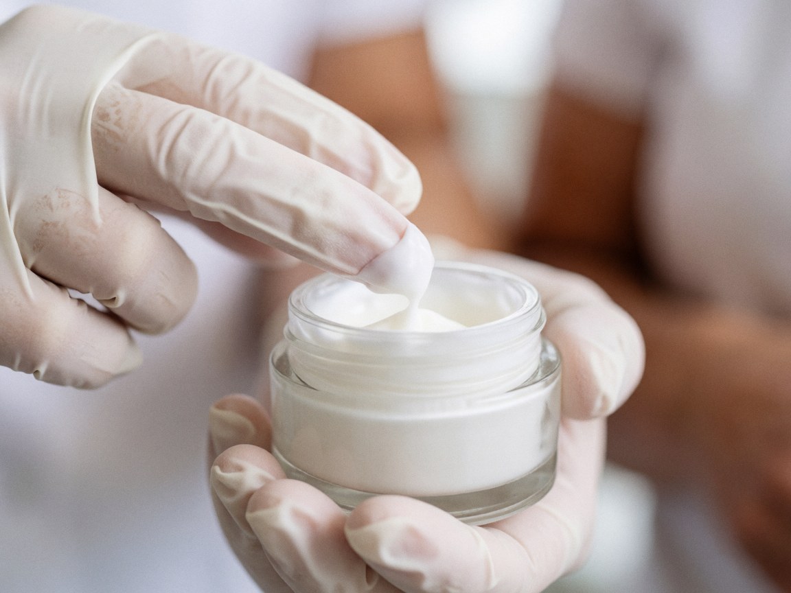 Close-up of gloved hands taking a portion of moisturizing cream from a glass jar against a clean, professional blurred background
