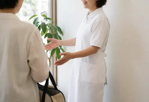 A therapist in white with open hands welcomes a client carrying a bag near plants and natural light at a health center entrance.