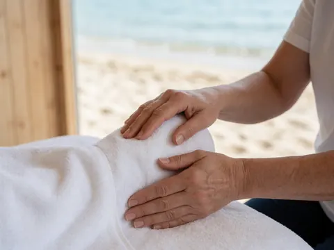 Therapist's hands performing a therapeutic massage on a foot covered with a white towel, set against a background of sea and light wood, creating a relaxing and professional atmosphere.