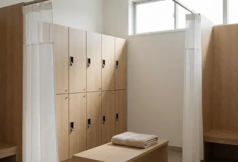 A clean and private dressing room featuring wooden closets, white curtains, and a folded towel on a bench, illuminated by natural light, ideal for therapeutic massage settings.
