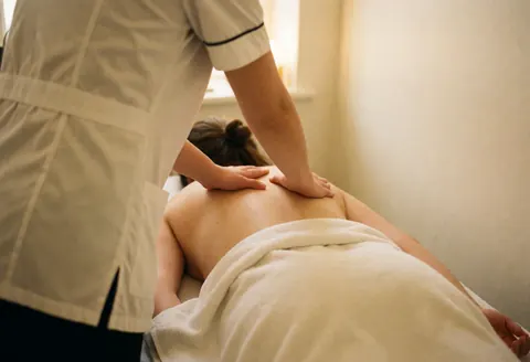 A therapist in a white uniform applies a back massage to a client covered with a white cloth in a warm, softly lit room.