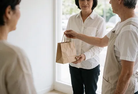 A therapist hands a paper bag to a client in a bright room. Both figures are blurred or facing away, conveying calm professionalism and attentive service.