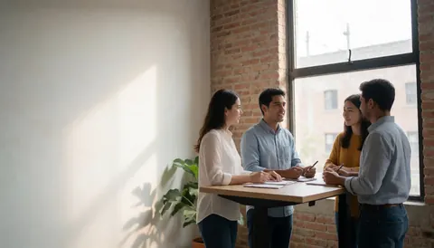 A bright modern office space with exposed brick walls, natural light, and professionals collaborating at shared desks with laptops and casual attire