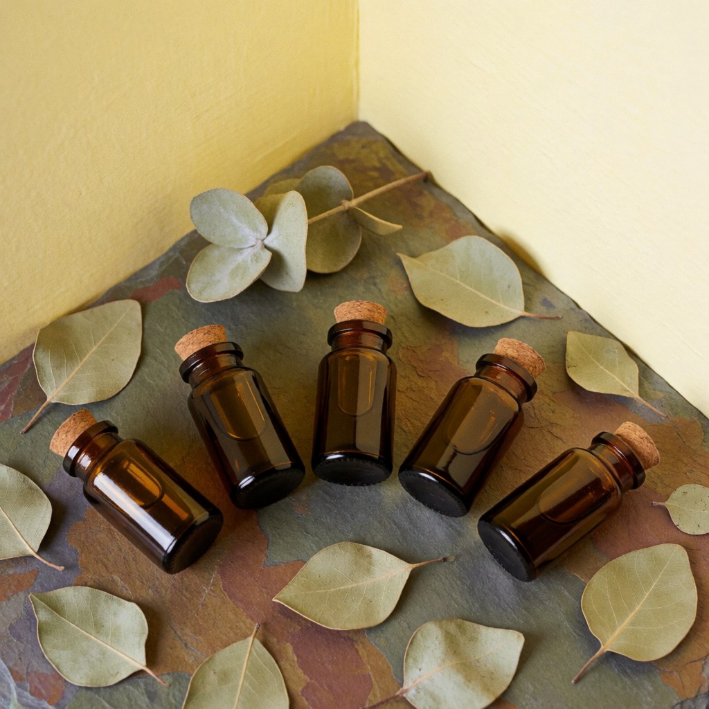 Top-down view of small glass essential oil bottles arranged with dried leaves and a stone on a light surface for therapeutic massage products