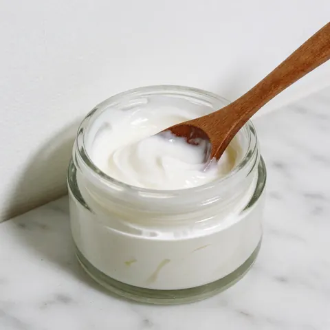 White therapeutic cream in a glass jar with a wooden spoon on a white marble surface, ideal for massages and skin care products.